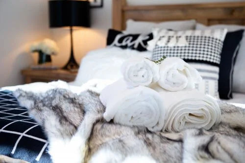 Stack of white towels on a bed with decorative pillows and a lamp in the background
