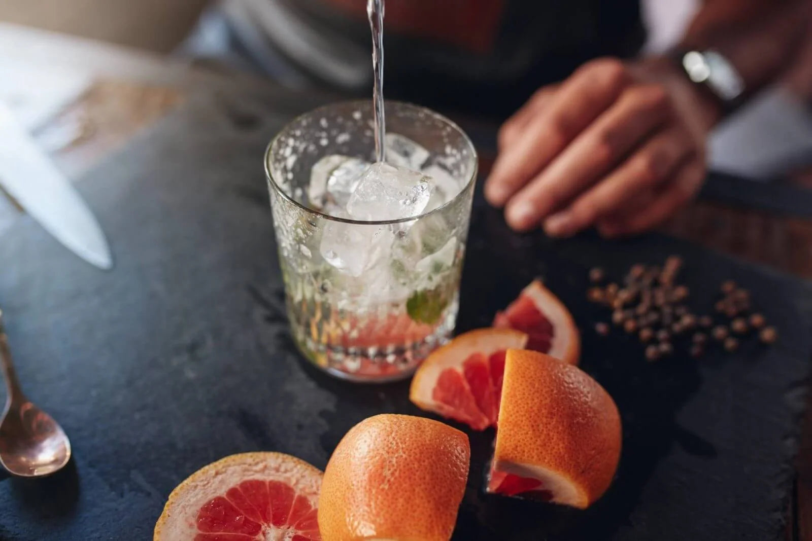 Person pouring a drink into a glass with ice, surrounded by sliced grapefruits on a dark surface.