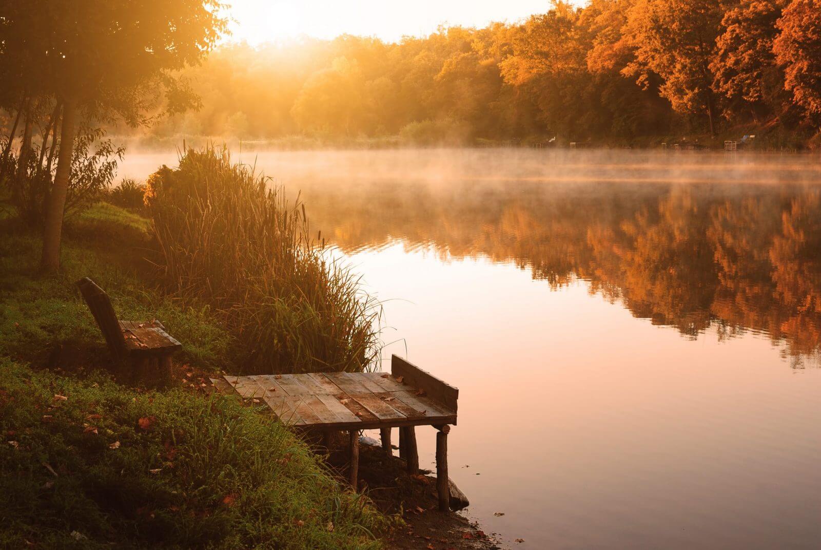 Peaceful lake scene with a wooden dock at sunrise or sunset, surrounded by trees.